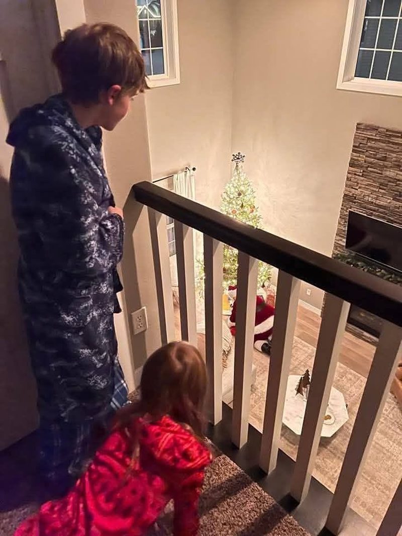 Two children in pajamas peeking over the stair banister at Santa placing gifts under the tree
